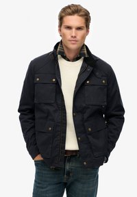 Marineblauw jack van duurzame stof met vier voorkralen, gouden drukknoopsluitingen en een rits, gelaagd over een crème sweater.