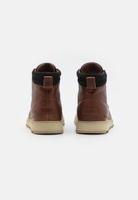 Des bottes ankle en cuir marron présentent un col en fourrure noire, un bout rond et une semelle en caoutchouc beige, avec une étiquette de logo au talon.