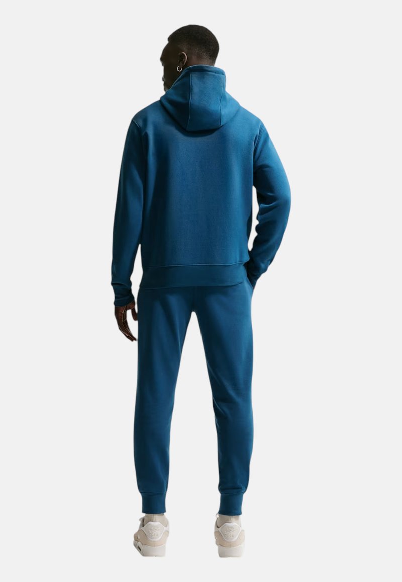 Homme portant un sweat à capuche bleu et un pantalon de jogging assorti, debout avec une main dans la poche, tourné de dos, sur un fond clair uni.