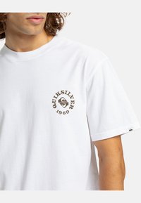 Biela bavlnená tričko s okrúhlym výstrihom, s hnedým kruhovým logom a textom "Quiksilver 1969" na ľavej hrudi. Krátke rukávy.