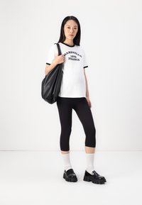 Hvit t-skjorte med svarte ringer og tekst, svarte leggings, kraftige svarte sko, og en strukturert svart veske. Modellen står mot en ensfarget bakgrunn.