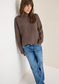 Brązowy sweter o fakturowanym splocie z wysokim kołnierzem, zestawiony z jasnoniebieskimi szerokimi dżinsami o klasycznym fasonie z pięcioma kieszeniami.