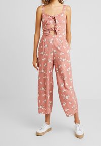 Frau in einem pinken, blumigen ärmellosen Jumpsuit mit zwei vorderen Knoten und weißen Plateauschuhen, die vor einem neutralen Hintergrund steht.