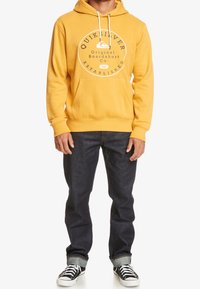 Felpa con cappuccio gialla e logo bianco, con tasca frontale e lacci; abbinata a jeans blu scuro e sneakers nere e bianche.
