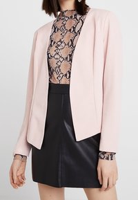 Femme portant un blazer rose clair sur un haut à imprimé serpent rentré dans une mini-jupe en cuir noire, debout devant un fond blanc.