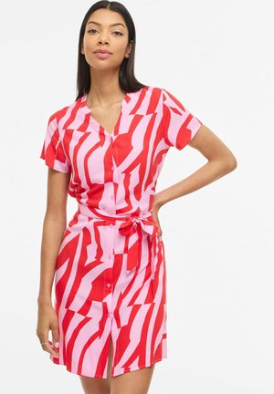 Kobieta w różowej sukience na guziki z krótkim rękawem, ozdobionej odważnymi czerwonymi, abstrakcyjnymi paskami i paskiem wiązanym w talii, stoi z jedną ręką na biodrze.