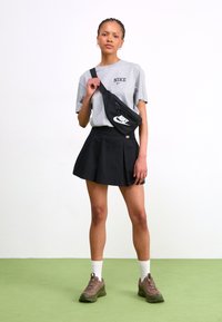 Czarna plisowana spódnica, szara koszulka Nike, czarna nerka z białym logo, brązowe sneakersy, białe skarpetki, warkocze. Prosty, sportowy strój.