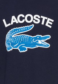 Modrý krokodýl s bílými skvrnami a otevřenou tlamou pod tučným bílým textem "LACOSTE" na tmavomodrém textilním pozadí.