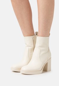 Bottes blanches en cuir avec un talon bloc haut, dotées d'une fermeture éclair latérale et d'une texture lisse. Conçues avec une esthétique minimaliste.