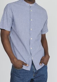 Lyhythihainen nappipaita sinivalkoisilla pystyraitoja, jossa on kauluksessa bändityyli ja kevyt kangas, päällä henkilö, jolla on kädet taskuissa.