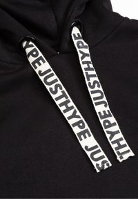 Sweat à capuche noir avec des cordons blancs arborant le texte noir répété « JUSTHYPE », enfilé à travers des œillets métalliques noirs.