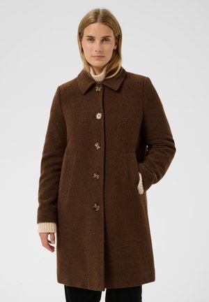 Femme portant un manteau en laine marron boutonné par-dessus un pull beige, une main dans la poche, debout devant un fond uni.