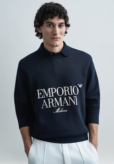 Sweat-shirt bleu marine avec col, affichant le texte "EMPORIO ARMANI" en couleur crème et un petit patch logo. Texture douce, poignets et ourlet côtelés.