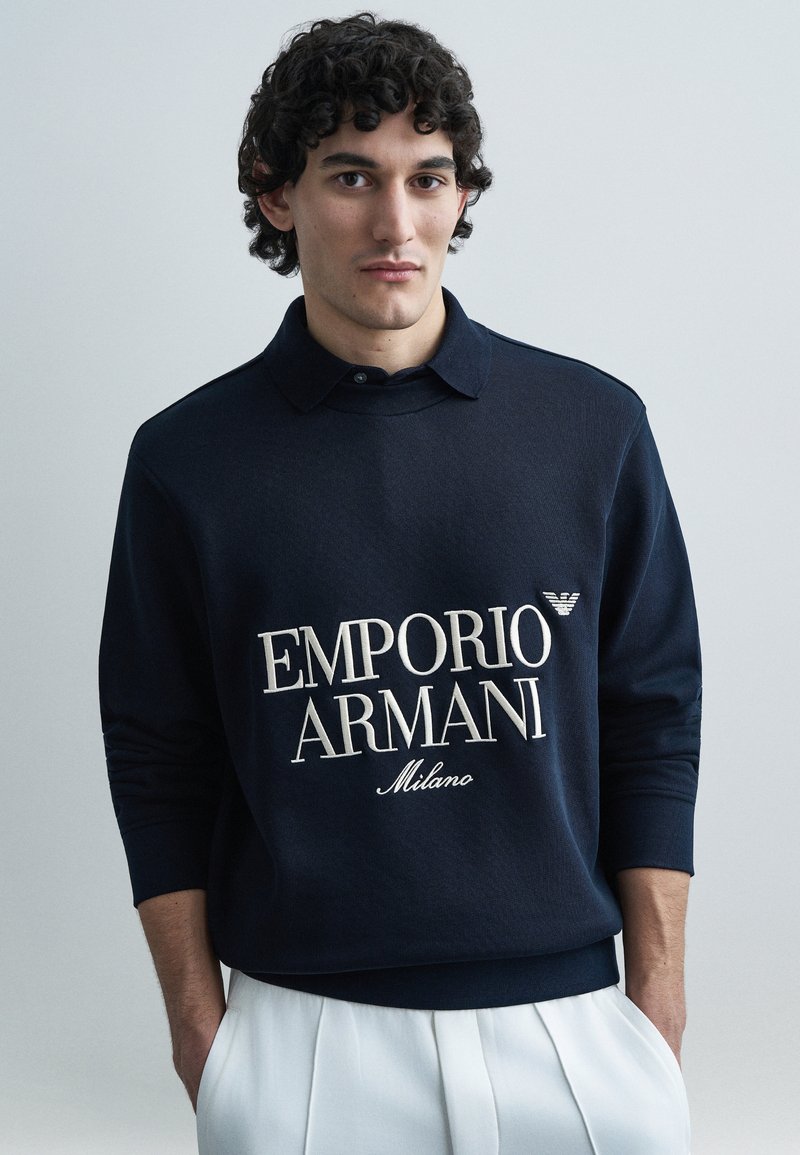 Granatowa bluza z kołnierzem, z napisem "EMPORIO ARMANI" w kolorze kremowym oraz małą naszywką z logo. Miękka tekstura, ściągacze na mankietach i u dołu.