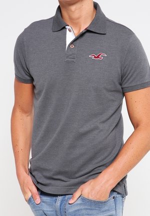 Mężczyzna w ciemnoszarej koszulce polo z małym czerwonym logo na piersi, w jasnoniebieskich dżinsach, ręce w kieszeniach.