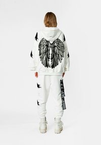 Sudadera con capucha blanca con diseños de alas negras en la espalda y detalles desgastados, combinada con pantalones deportivos a juego y botas blancas.