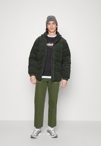 Dark green puffer jacket with elastic cuffs, paired with olive green pants and a black sweater. Grey beanie and patterned sneakers complete the look.