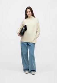 Džemper od kremasto bijelog, mekanog materijala s visokim ovratnikom, uparen s ležernim plavim trapericama i bež cipelama. U ruci drži crnu torbicu.