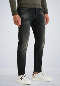 Jeans ajustados de mezclilla oscura con un desgaste sutil, cinco bolsillos y cierre de botón. Combinados con zapatillas blancas y un sudadera verde olivo.