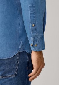 Lichtblauwe spijkerblouse met knoopmanchetten, voorzien van een puntige kraag en een ronde zoom. Draagt over blauwe jeans, wat een casual uitstraling geeft.