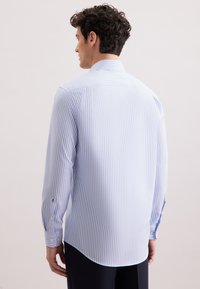 Homme portant une chemise à rayures verticales bleu clair et blanche à manches longues, vu de dos sur un fond uni.