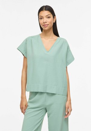 Kobieta z długimi ciemnymi włosami, ubrana w luźny, jasnozielony top z krótkim rękawem i dekoltem w serek oraz pasujące spodnie, stojąca na białym tle.