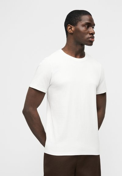 Άνδρας που στέκεται με τα χέρια πίσω από την πλάτη, φοράει απλό λευκό κοντομάνικο μπλουζάκι και σκουρόχρωμο παντελόνι, κοιτάζοντας προς τα δεξιά μπροστά σε λευκό φόντο.