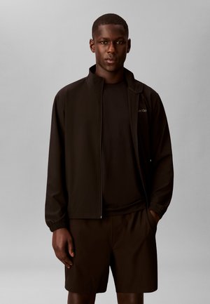 Homme portant une veste noire zippée, un t-shirt noir et un short noir, debout devant un fond gris uni.