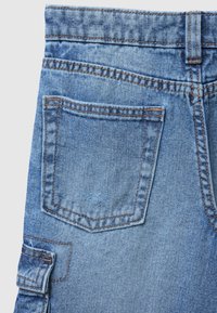 Tessuto denim di colore azzurro chiaro, con una tasca posteriore cucita di forma rettangolare e fili marroni a contrasto. Nessun motivo visibile.