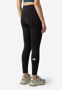 Leggings noires avec une taille haute, en matériau extensible, dotés d'un logo blanc sur la jambe inférieure. Portés avec des baskets grises et beiges.