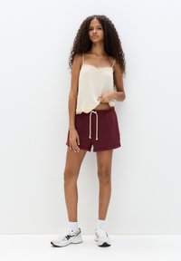 Krämfärgad kamsol med spetsdetaljer och knappar, tillsammans med vinröda shorts med snörning. Modellen bär vita sneakers med grå accenter.
