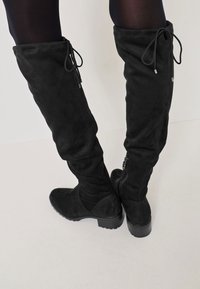 Botas negras hasta la rodilla con una suave textura de ante, punta redondeada, detalles de cordones en la parte trasera y un tacón grueso. Cierre lateral con cremallera visible.