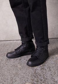 Osoba w czarnych butach wysokich i czarnych spodni cuffed stoi na teksturowanej betonowej podłodze.