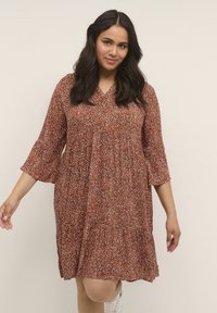 Braunes Blumenkleid mit V-Ausschnitt, drei Viertel langen Glockenärmeln und fließender Silhouette, verziert mit kleinen weißen und orangenen Mustern.
