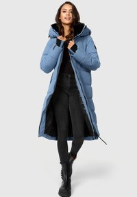 Abrigo acolchado azul con capucha, con un forro de piel sintética, de largo extendido, cierre de cremallera y detalles en negro, combinado con jeans negros y botas.