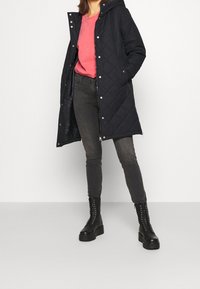 Abrigo de invierno acolchado negro con capucha, jeans ajustados grises y botas negras con cordones. El abrigo cuenta con botones a presión y un diseño largo y ajustado.