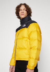 Puffjakke med sort overdel og klart gul underdel. Har en høj krave, quiltet tekstur og logo på brystet.