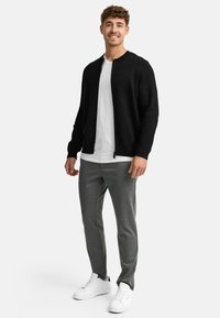 Chaleco negro con cremallera sobre una camiseta blanca, combinado con pantalones grises ajustados y zapatillas blancas. Tejido suave con un diseño minimalista.