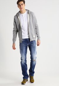 Grå hettejakke med glidelås og snorer, hvit henley-skjorte, slitte blå jeans og olivengrønne sneakers med hvit såle.