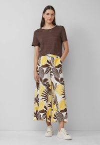 T-shirt court marron associé à un pantalon large avec un motif de feuilles jaunes et marron. Le mannequin porte des baskets blanches.