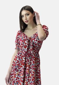 Vestido de estampado floral con mangas abullonadas, flores rojas, azules y naranjas sobre un fondo blanco. Presenta un lazo en la parte delantera y una cintura fruncida.