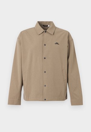 Beige nappikoru takki pitkillä hihoilla, kauluksella, sivutaskuilla ja pienellä mustalla geometrisella logolla vasemmassa rinnassa.