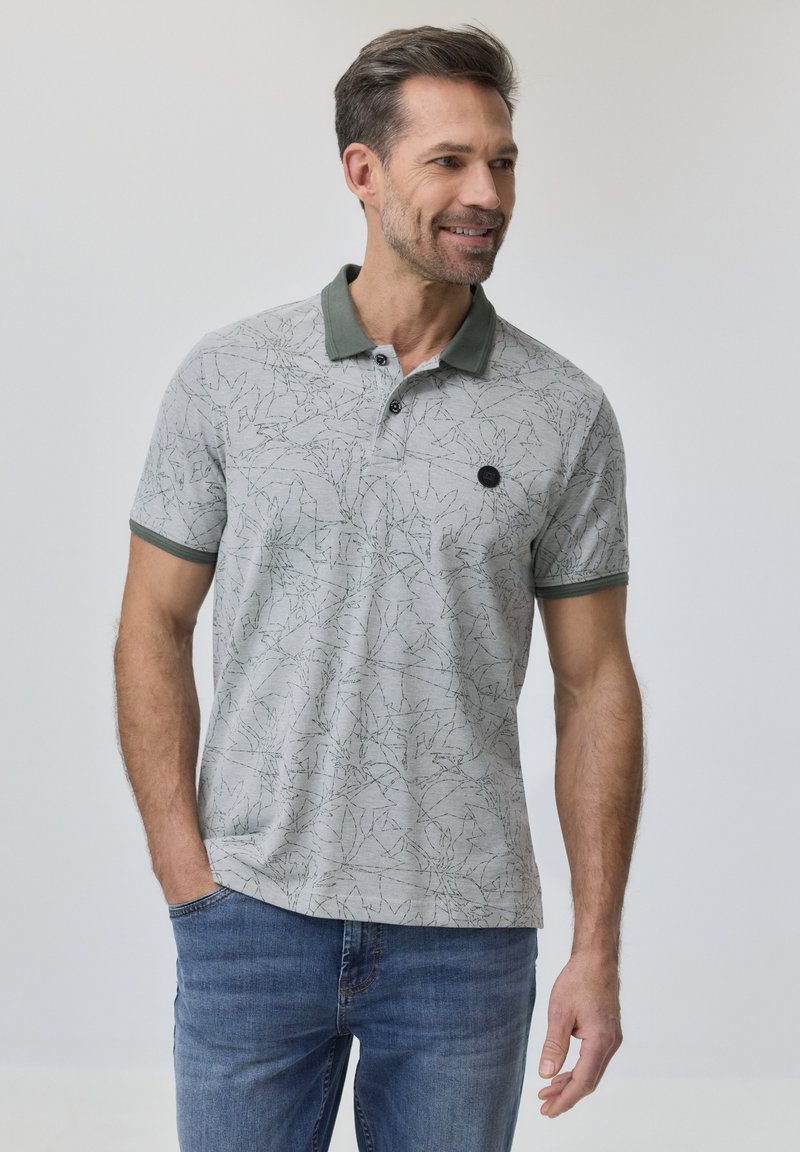 Homme portant un polo gris clair à motifs avec un col et des bordures verts, debout avec une main dans la poche d'un jean bleu, souriant et regardant vers la droite.
