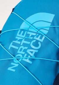 Mochila de tecido azul com um logótipo em destaque, detalhes em corda elástica e superfície texturizada. O design enfatiza a durabilidade e a funcionalidade.