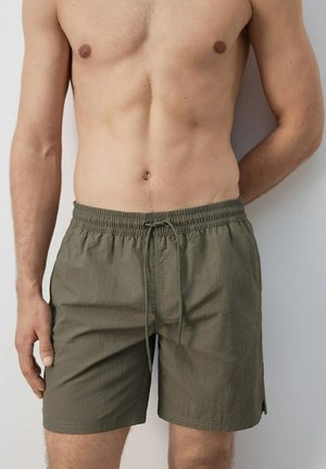 Shirtless man wearing olive green drawstring shorts, standing with one arm behind his back against a plain background.