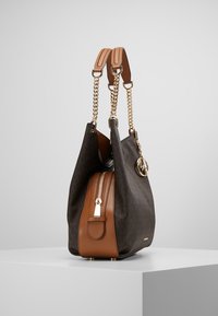 Sac en tissu texturé marron avec une chaîne dorée et des accents en cuir tan. Il a une fermeture éclair et un charme de logo. Vue de côté.