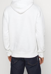 Sudadera blanca con capucha que presenta una textura suave, puños y dobladillo acanalados, y un diseño liso sin logotipos ni patrones visibles.