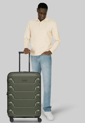 Hombre con camisa beige de manga larga y vaqueros sosteniendo el asa de una maleta grande verde de carcasa rígida con cuatro ruedas sobre fondo gris.