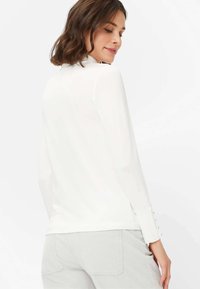 Hvid langærmet top med høj hals og knapper ved håndleddene. Glat tekstur, tætsiddende design og en subtil glans.