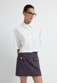 Witte blouse met korte mouwen en knoopsluiting, versierd met parelachtige details, gecombineerd met een aansluitende donkerblauwe en rode textuurrijkrok met gouden knopen.